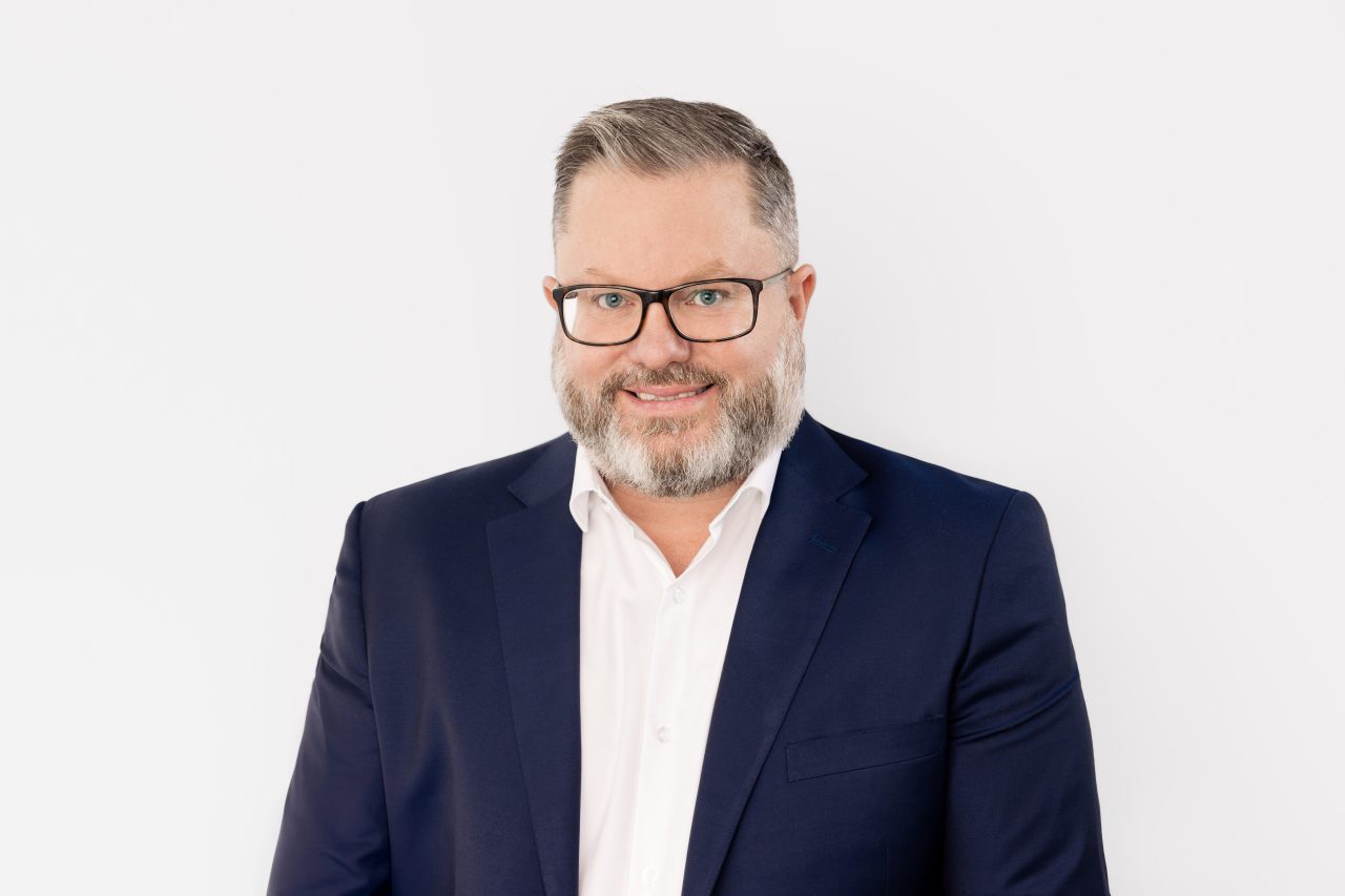 Professional studio headshot of a spokesperson wearing glasses, a navy suit and white shirt against a light grey background.