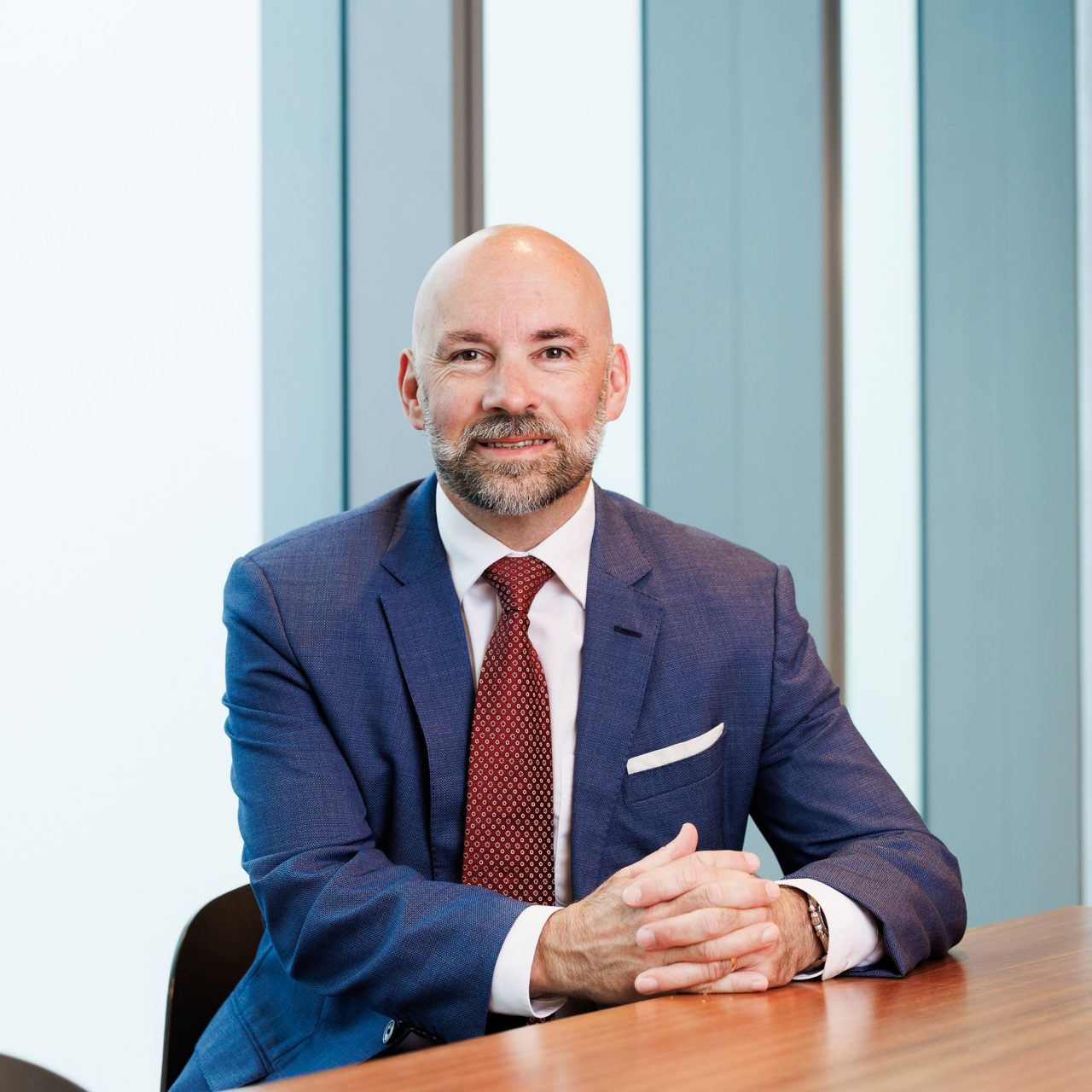 Person seated at a meeting table in an office setting, wearing a blue suit, white shirt and red tie.