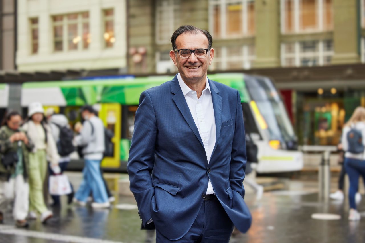 Michael Saadie in a blue suit standing with hands in pockets on street with tram behind